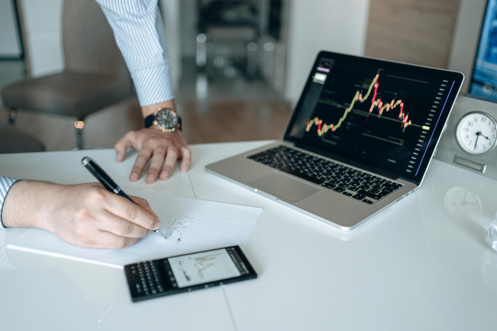 An angled, high-angle shot of a white office desk where a person is writing on a sheet of paper with a pen. Next to them, a laptop displays a colorful stock market candlestick chart, and a classic Blackberry smartphone sits nearby, also showing a financial graph.