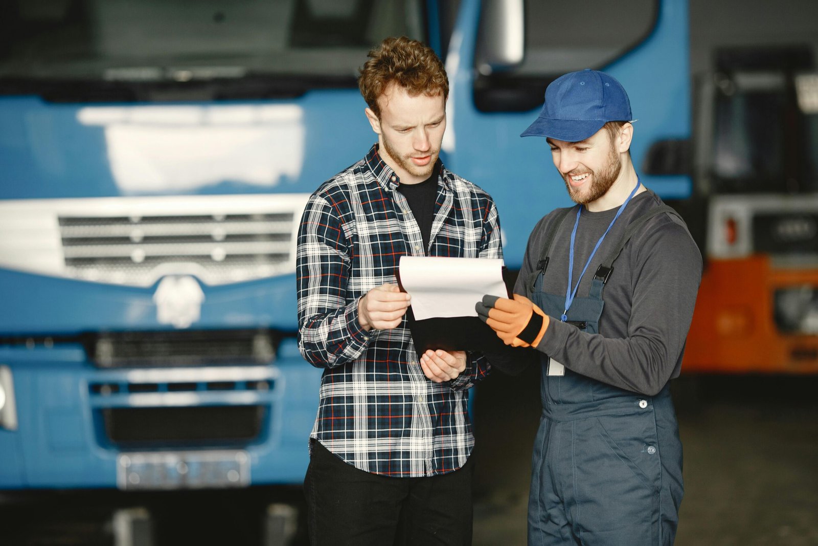 A man in a plaid shirt and a mechanic in blue overalls and a cap stand together in a garage, reviewing a document on a clipboard in front of a large blue truck.