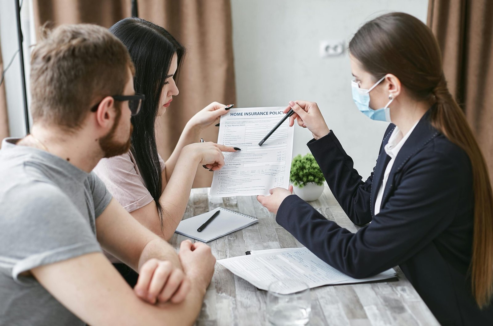 An insurance agent wearing a blue surgical mask and a navy blazer points to a "Home Insurance Policy" document with a pen while a young couple reviews it across a wooden desk in an office setting.