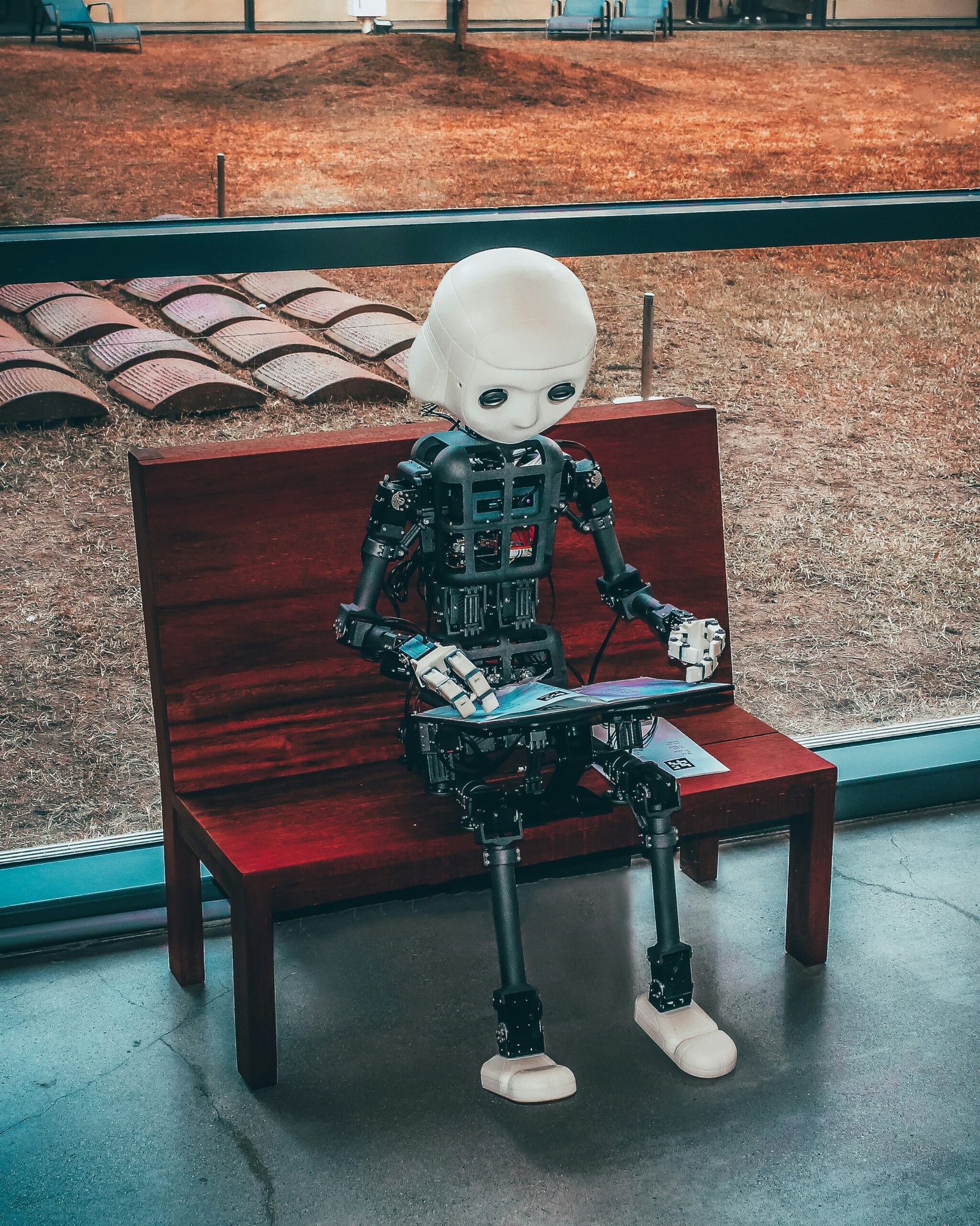 An iCub humanoid research robot with a white face and exposed mechanical body sitting on a red wooden bench while holding a blue tablet, positioned in front of a large window overlooking a yard.