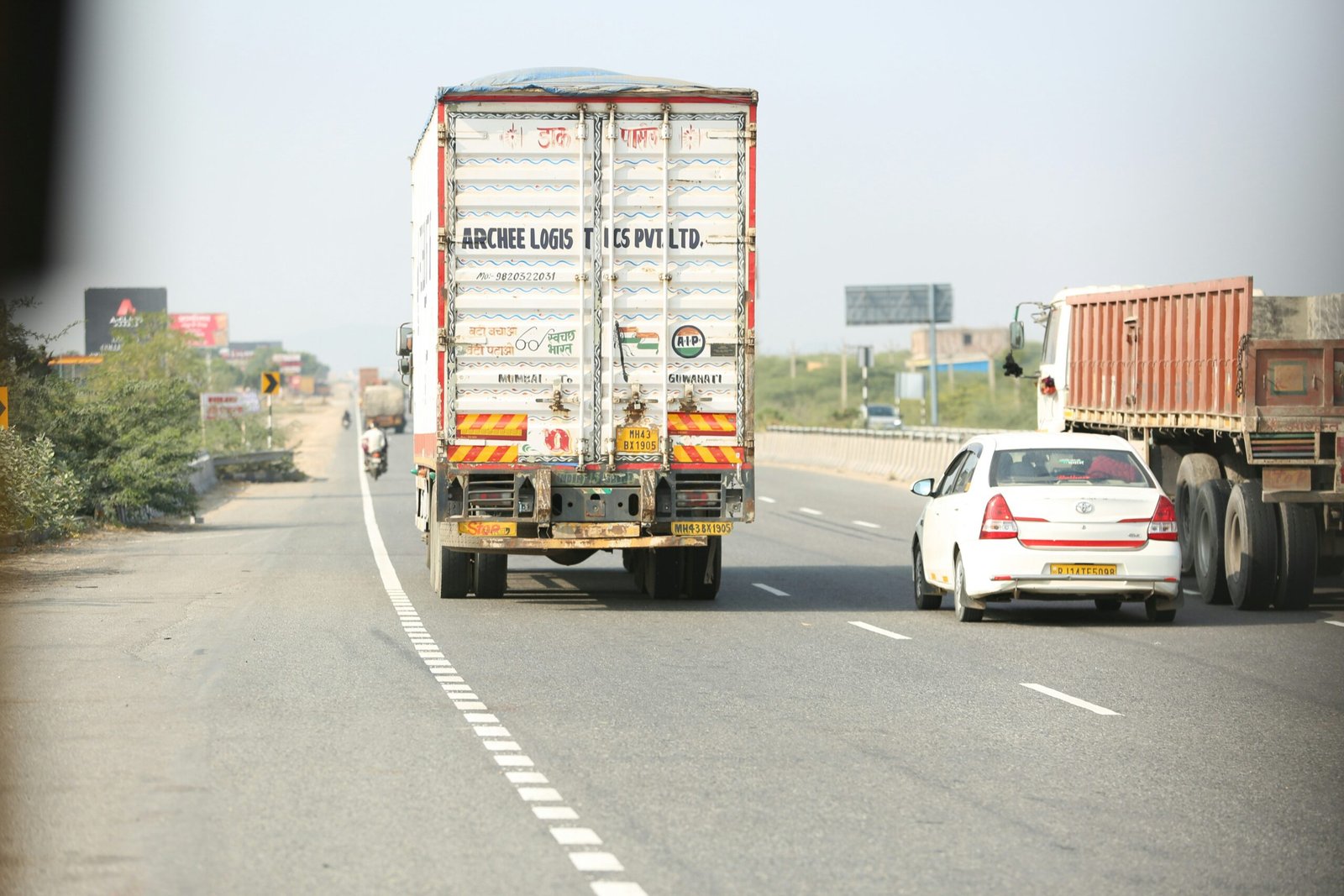 A rear view of a white commercial truck from Archee Logistics Pvt Ltd. driving on a multi-lane paved highway in India, alongside a white sedan and a large brown flatbed truck under a bright, clear sky.