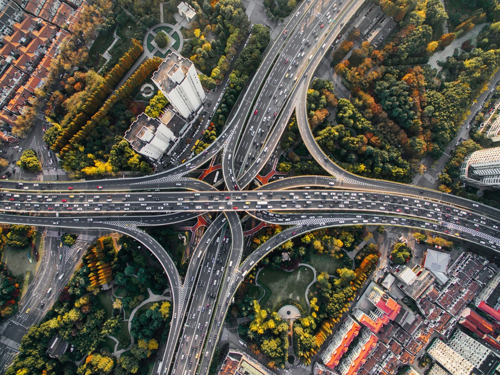 Aerial top-down view of a complex multi-level highway interchange with heavy traffic, surrounded by green and yellow trees and urban buildings.