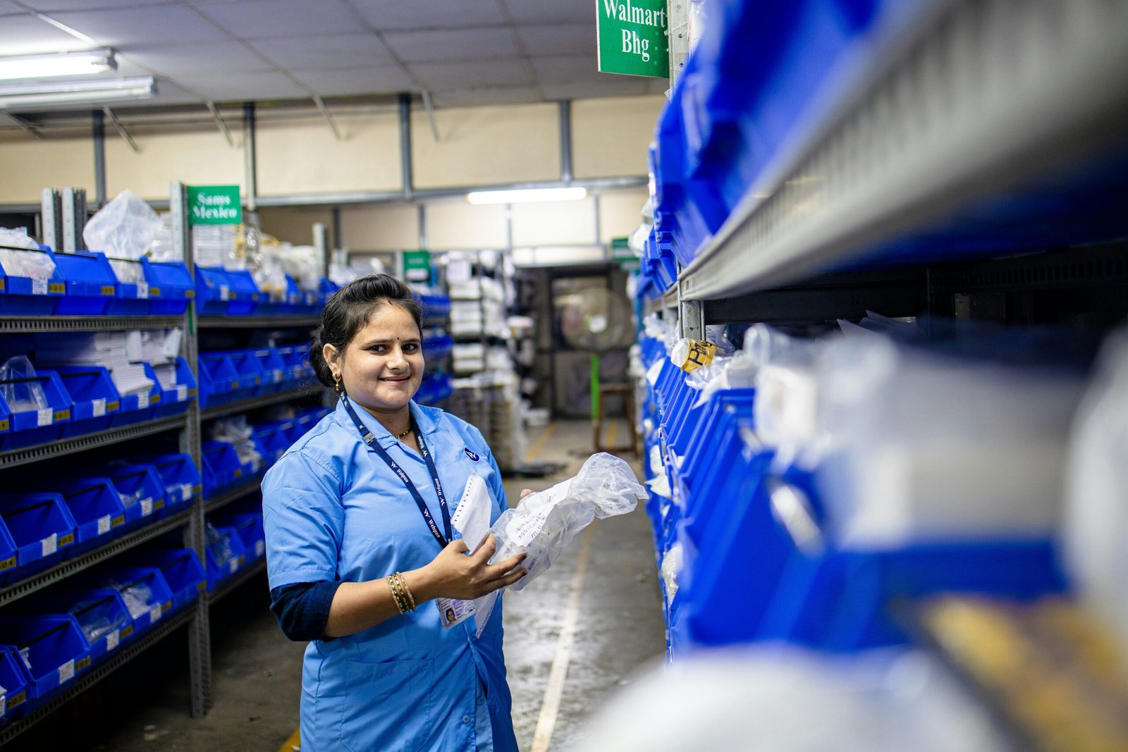 A smiling female warehouse associate in a blue uniform stands in an aisle lined with blue storage bins, holding a packaged item.