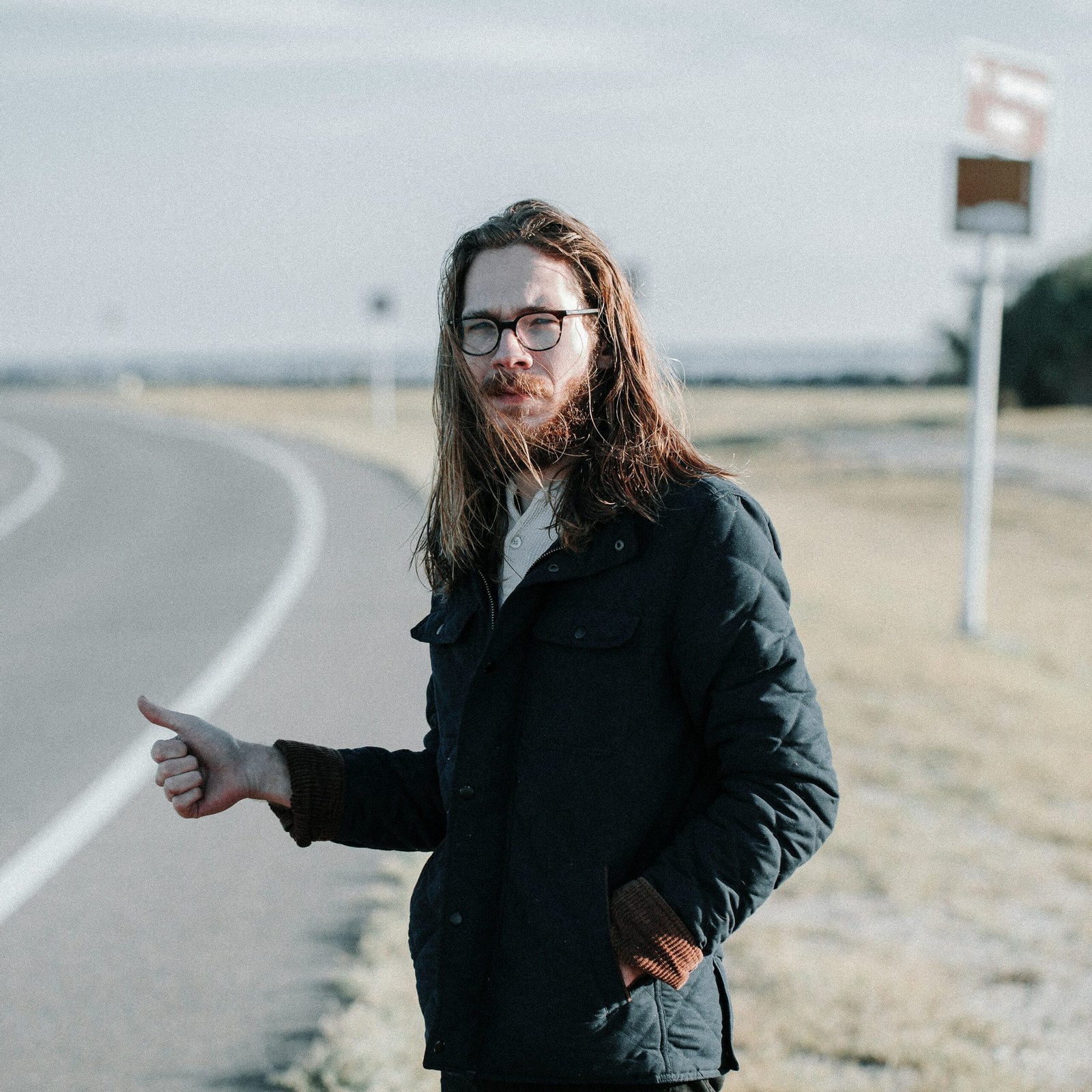 A portrait of a man with long brown hair, a beard, and round glasses, standing on the edge of a winding paved road. He is wearing a dark, quilted jacket over a light-colored button-up shirt and is holding his thumb out in a hitchhiking gesture. The background shows a flat, dry landscape under a pale, overcast sky, with a blurred road sign visible in the distance.