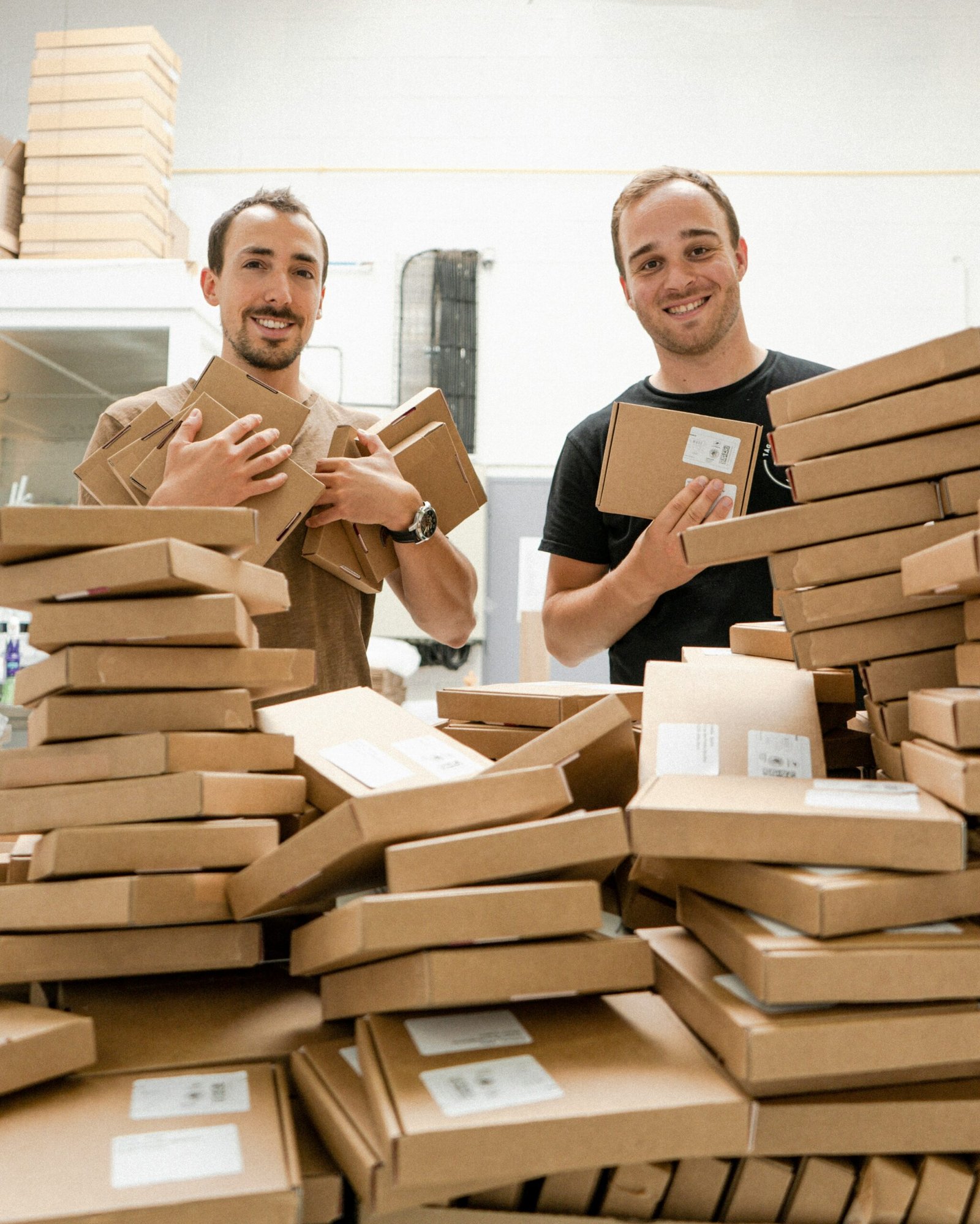 Two smiling men standing in a warehouse, holding and surrounded by numerous stacks of small brown cardboard shipping boxes ready for delivery.