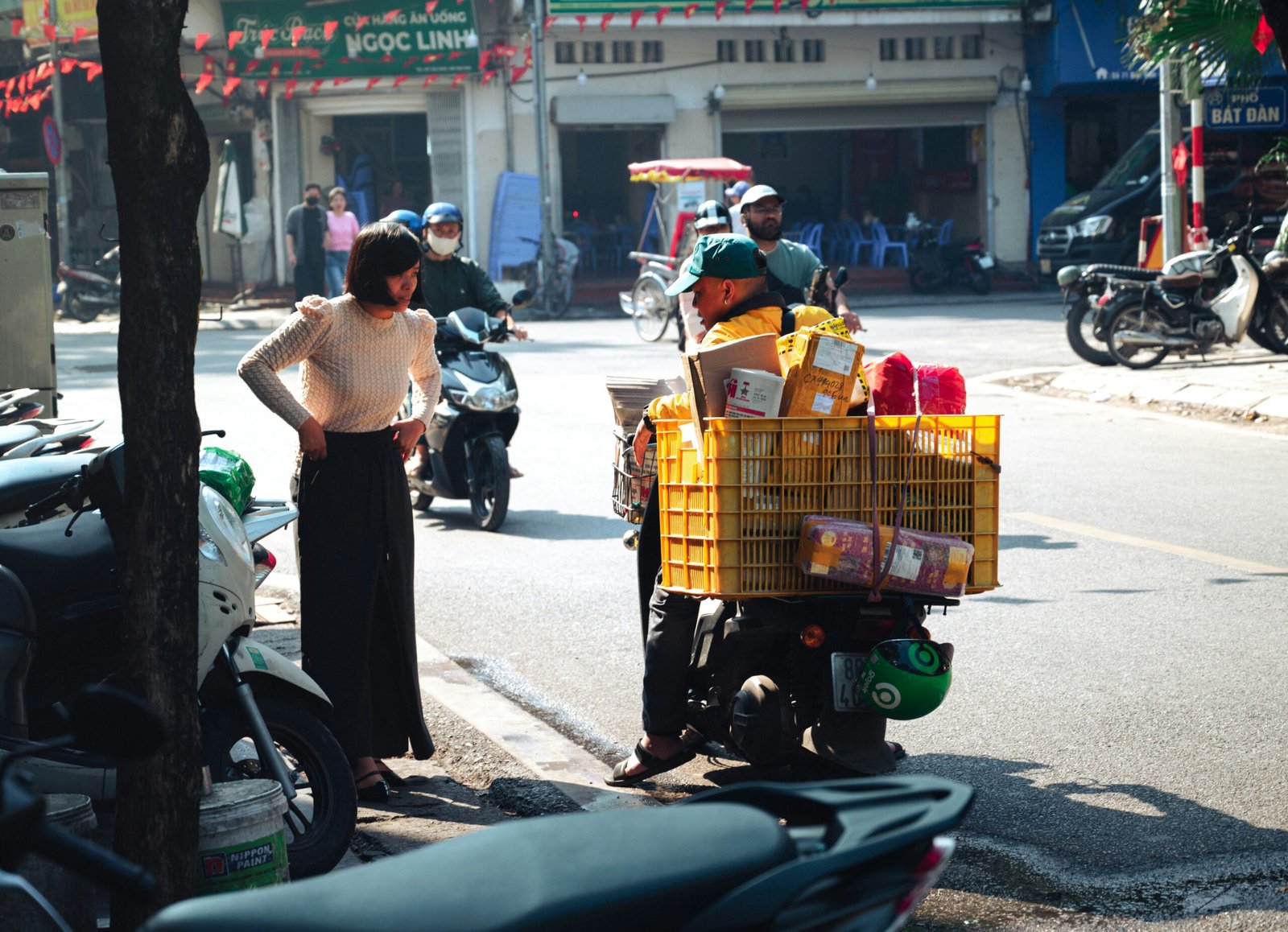 A street scene in Vietnam featuring a delivery person on a motorbike heavily loaded with a yellow plastic crate full of packages. They are interacting with a woman standing on the sidewalk wearing a light-colored knit sweater and dark skirt. In the background, other motorbikes navigate the sunlit street lined with local shops and buildings.