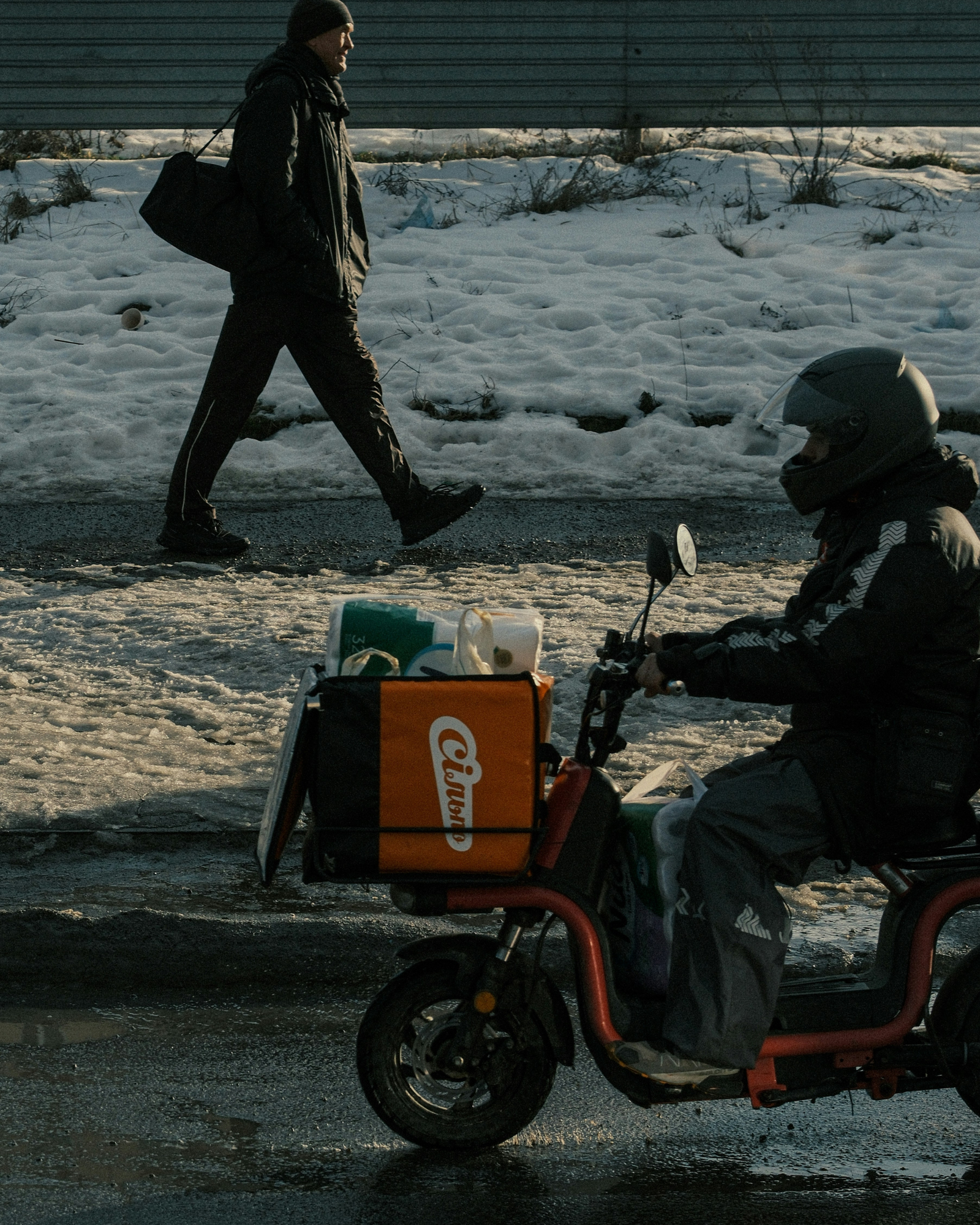 A delivery rider on an orange electric scooter with a large cargo box in the foreground, while a man in dark winter clothing walks along a snowy roadside in the background.
