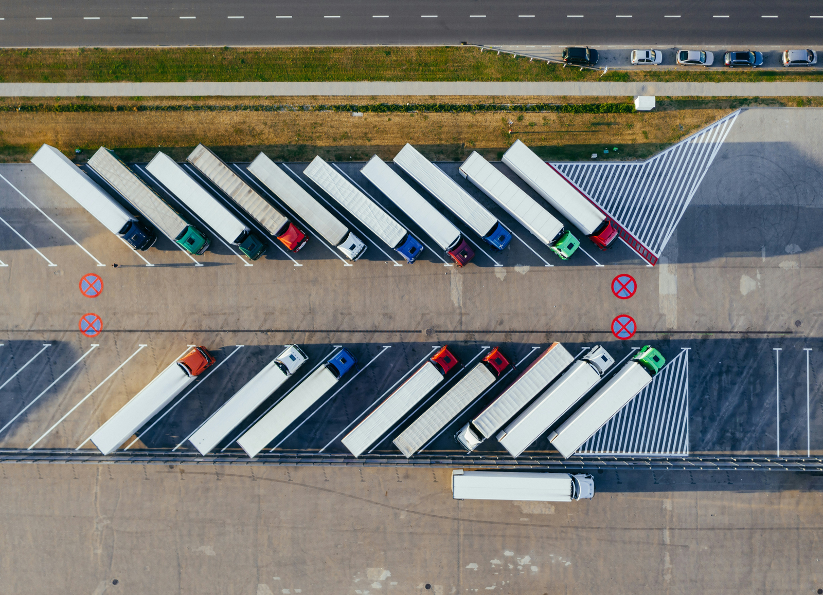 An overhead drone shot of a paved storage yard filled with various utility vehicles. The majority of the fleet consists of heavy-duty yellow dump trucks, flatbeds, and service trucks arranged in organized rows. A small section in the bottom left features several white passenger cars and a white shuttle bus parked together. Long, sharp shadows from the late afternoon or early morning sun stretch across the cracked asphalt between the vehicles.
